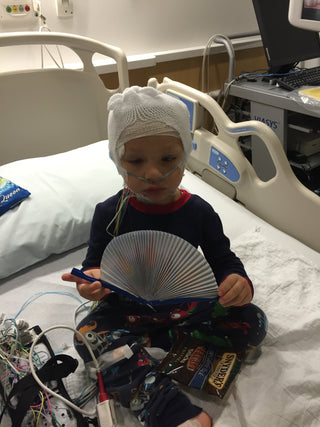 Child in pediatric hospital bed wearing medical head wrap and oxygen tube, holding a toy fan, with monitoring wires and equipment—capturing pediatric neurology care and hospital stay.
