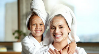 Smiling mother and daughter wearing white bathrobes and towels wrapped on their heads, with facial masks applied, enjoying a fun and relaxing skincare moment together.