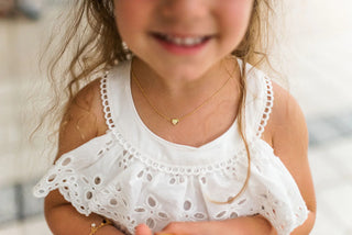Close-up of a child wearing a white lace top and solid heart pendant necklace with a blurred background