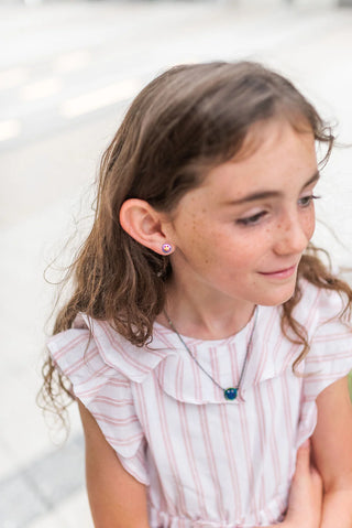 Young girl wearing a pink striped dress with ruffled details, a stainless steel rainbow metallic smiley necklace and matching smiley face earrings sitting against a white background.