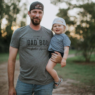 Man holding a child outdoors with a casual background in a shirt that says It's Not a Dad Bod its a Father Figure.