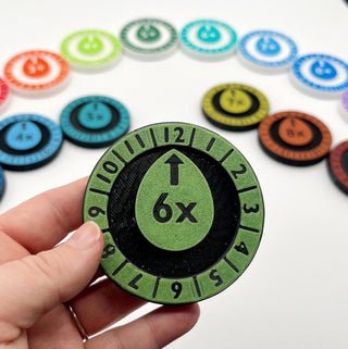 Hand holding a green game token with numbers and symbols, surrounded by other similar tokens on a white background.