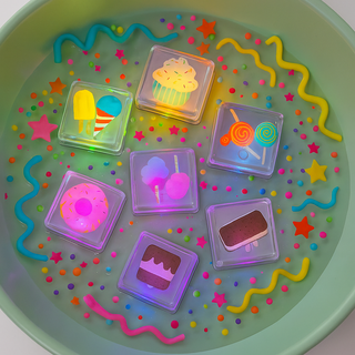 Colorful light up cubes decorated like ice cream, donuts, and other treats floating in a green bowl with decorative elements.