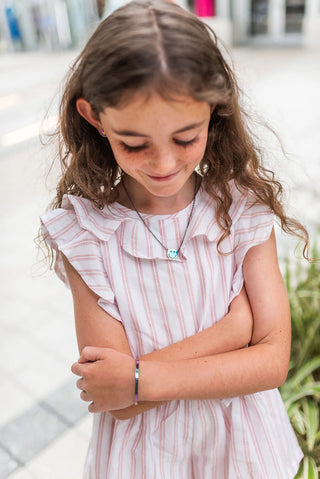 Young girl wearing a pink striped dress with ruffled collar and stainless steel metallic rainbow smiley face pendant necklace, standing outdoors.