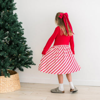 Child in a red top and striped skirt standing next to a Christmas tree indoors.