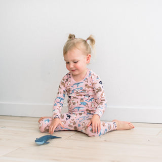 Child wearing a pink pajama with animal prints sitting on a light wood floor.