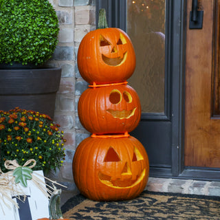 Three stacked carved pumpkins in front of a door with decorative plants.