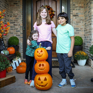 Three children standing in front of a house with three stacked jack-o'-lanterns on a porch.