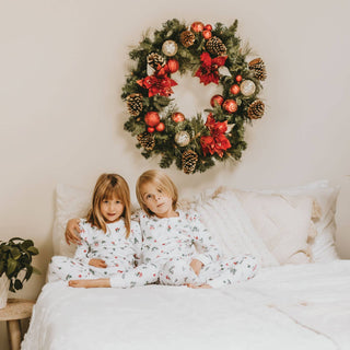 Two children in matching pajamas sitting on a bed with a decorated Christmas wreath above them.