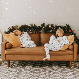 Two children in pajamas sitting on a brown couch with Christmas decorations in the background.